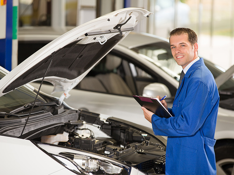 Summer road trip vehicle inspection at Homan GMC of Ripon being done by a bright blue-coated certified professional mechanic wearing a tie, as he pauses to smile at us, pen checking off the last task he accomplished on the white vehicle's open-hooded engine in the service center